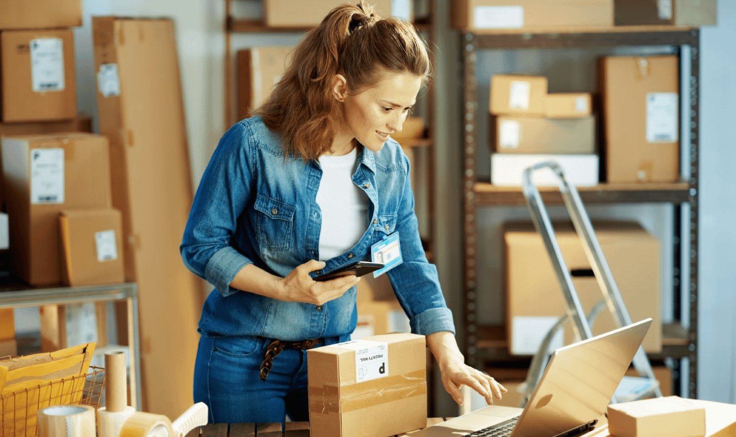 Woman checks laptop while stood in warehouse full of boxes